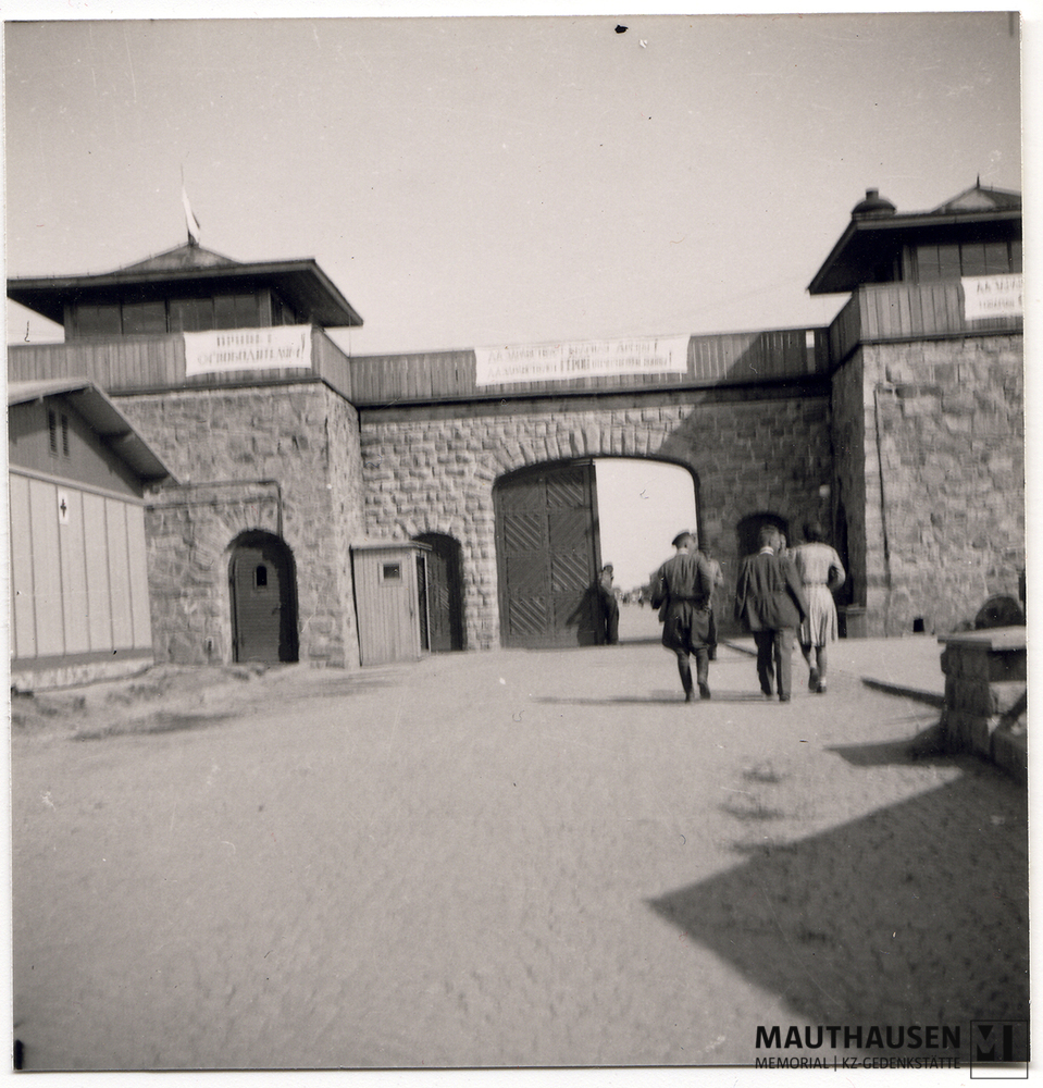 MM_4_7_5_0019.jpg; 4.7.5.0019; Befreites KZ Mauthausen: Blick auf das Jourhaus mit geöffnetem Lagertor von außen Richtung Appellplatz, im Vordergrund eine Personengruppe. ;
